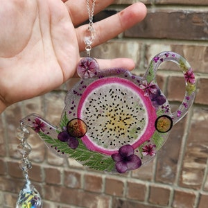 May include: A clear resin teapot ornament with a dragon fruit slice, dried flowers, and glitter. The teapot is hanging from a silver chain with a crystal pendant.