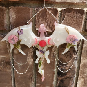May include: A white dragon silhouette with pink and red accents, decorated with pressed flowers and hanging from silver chains. The dragon has a moon and star design on its wings.