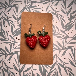 Peut inclure: Une paire de boucles d'oreilles en forme de cœur rouge avec des tiges vertes et des crochets argentés. Les boucles d'oreilles sont présentées sur une carte marron.