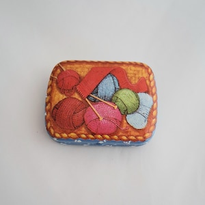 May include: A rectangular tin with a blue border and a decorative illustration of yarn balls, knitting needles, and a red scarf. The tin has a gold-colored background with a woven texture.