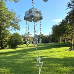 May include: A whimsical wind chime featuring a cloud design with iridescent orbs and dangling chains. The chains support clear, faceted beads and a clear lightning bolt charm. The wind chime is suspended against a backdrop of a green lawn and blue sky.
