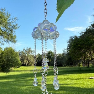 May include: A hanging suncatcher with a cloud-shaped top made of iridescent glass beads. Below the cloud are chains with clear crystal accents and a sun-shaped charm. The suncatcher hangs from a silver chain against a blue sky and green grass.