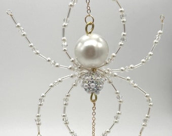 May include: A decorative spider ornament, made with clear beads, faux pearls, and gold-coloured chain. The spider's body features a large white pearl, with a cluster of smaller pearls and crystals. A dangling crystal flower hangs below.