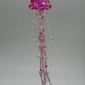May include: A handmade beaded jellyfish ornament. The top is a cluster of pink and magenta beads, with clear and pink beaded strands hanging below. The ornament is suspended by a silver chain.