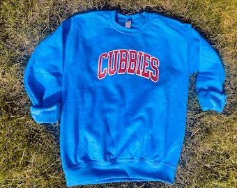May include: A blue crewneck sweatshirt with the word "CUBBIES" arched across the chest in red and white lettering. The sweatshirt has cuffed sleeves and a ribbed hem. The garment is displayed on a bed of green grass.