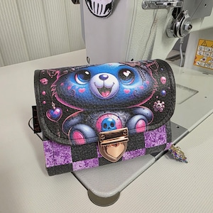 Pode incluir: Uma carteira preta e roxa com um desenho de urso de desenho animado. O urso é azul com detalhes em rosa e uma caveira no peito. A carteira tem um fecho em forma de coração e um padrão xadrez na parte inferior.