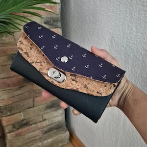 Peut inclure: Un portefeuille bleu marine avec un design en liège et cuir noir. Le rabat présente un motif d'ancres blanches. Le portefeuille a une fermeture argentée.