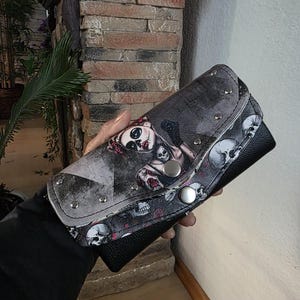 Könnte beinhalten: Eine schwarze Geldbörse mit einer Klappe, die einen Grafikdruck einer stilisierten Figur mit Totenkopf-Make-up zeigt, umgeben von einem Muster aus Totenköpfen und roten Rosen. Die Klappe ist mit zwei silberfarbenen Metalldruckknöpfen befestigt.