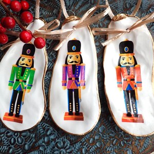 Puede incluir: Tres adornos de concha de ostra blanca con ribetes dorados, cada uno con un cascanueces de un color diferente. Los cascanueces llevan uniformes verde, morado y rojo con detalles dorados. Los adornos tienen un aspecto rústico y hecho a mano.