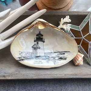 May include: A white seashell with a gold rim, painted with a black and white image of Brant Point Lighthouse in Nantucket, MA.