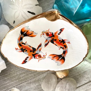 May include: A white seashell dish with a gold rim, featuring two orange and black koi fish painted on the surface.