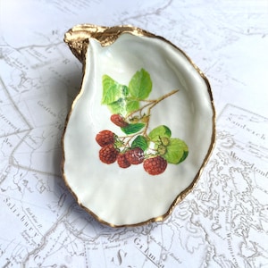 Puede incluir: Un plato decorativo de concha de ostra con borde dorado. El interior blanco presenta una ilustración de frambuesas rojas y hojas verdes. Una pieza de acento única y elegante.