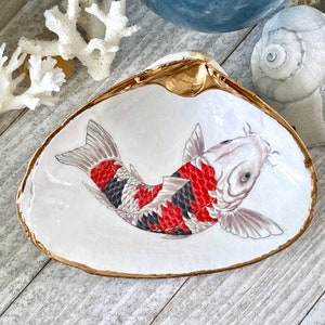 May include: A white seashell with a gold rim, featuring a hand-painted red, black, and white koi fish. The shell is a decorative dish or trinket.