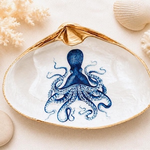 May include: A white clam shell dish featuring a detailed blue octopus illustration in the centre. The interior of the shell has a pearlescent finish, and the outer edges are trimmed with a metallic gold paint, highlighting the natural shell shape.