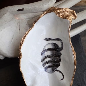 May include: A white ceramic oyster shell dish with a gold rim. The dish features a black and white illustration of a snake wrapped around an egg.