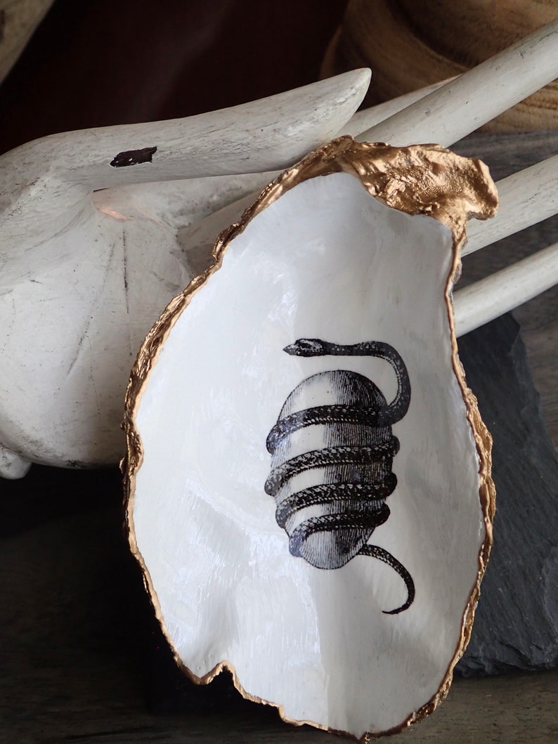 May include: A white ceramic oyster shell dish with a gold rim. The dish features a black and white illustration of a snake wrapped around an egg.
