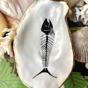 Puede incluir: Un plato de concha de ostra blanca con un borde pintado en oro y una ilustraci&oacute;n de esqueleto de pez pintada a mano en negro en el centro. La concha tiene un acabado liso y brillante y una forma natural irregular, resaltando el contraste entre la superficie blanca y el gr&aacute;fico esquel&eacute;tico oscuro.