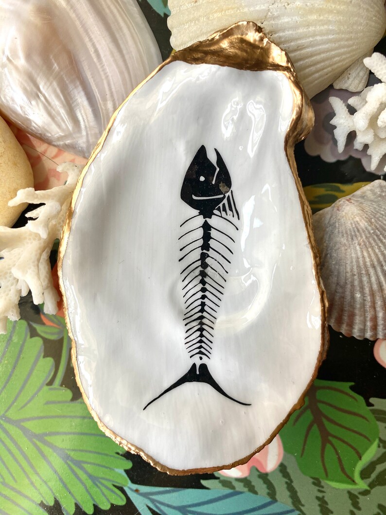 Puede incluir: Un plato de concha de ostra blanca con un borde pintado en oro y una ilustraci&oacute;n de esqueleto de pez pintada a mano en negro en el centro. La concha tiene un acabado liso y brillante y una forma natural irregular, resaltando el contraste entre la superficie blanca y el gr&aacute;fico esquel&eacute;tico oscuro.