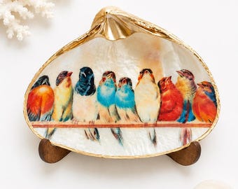 May include: A decorative clam shell dish featuring a painted row of nine colorful birds perched on a branch. The shell has a polished white interior with a metallic gold painted rim and is supported by a small dark brown wooden stand.