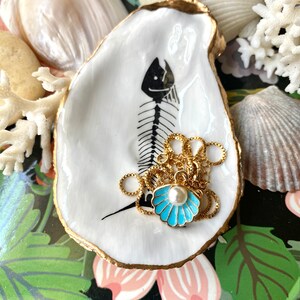 Puede incluir: Un plato de concha de ostra blanca con borde dorado y una ilustraci&oacute;n de esqueleto de pez en negro. En su interior descansa un collar de cadena dorada con un colgante de concha de esmalte azul claro y una perla blanca central.