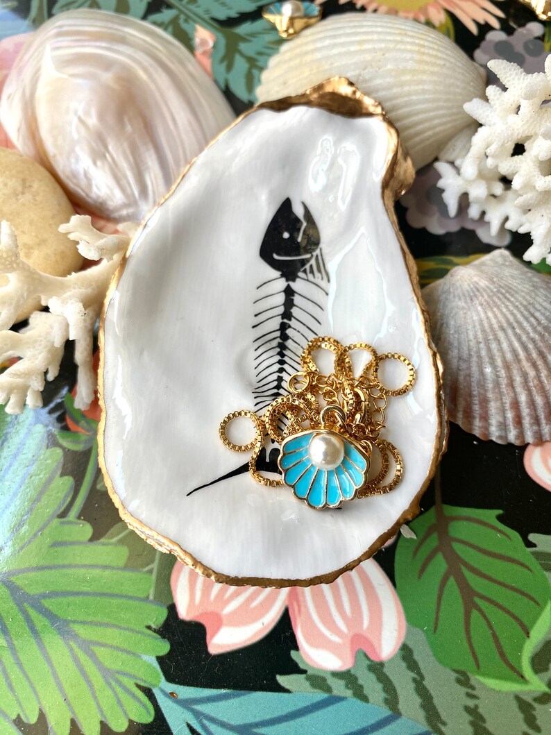 Puede incluir: Un plato de concha de ostra blanca con borde dorado y una ilustraci&oacute;n de esqueleto de pez en negro. En su interior descansa un collar de cadena dorada con un colgante de concha de esmalte azul claro y una perla blanca central.