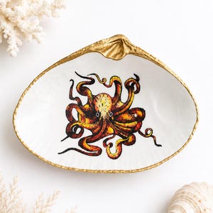 May include: A white clam shell dish featuring a hand-painted orange and yellow octopus illustration in the center. The edges of the shell are painted with a metallic gold finish, creating a decorative border around the white interior surface.