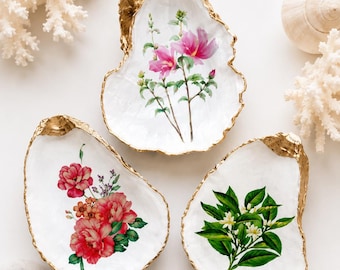May include: Three decorative oyster shells with gold-painted edges. The top shell features pink hibiscus flowers. The bottom left shell displays a cluster of red and pink peonies. The bottom right shell shows green leaves with small white flowers.