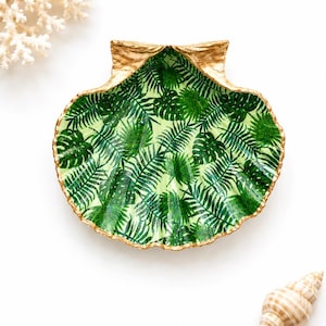 May include: A decorative scallop shell dish featuring a vibrant green botanical print of fern and monstera leaves on the interior. The edges and the hinge area of the shell are finished with a metallic gold paint, providing a distinct contrast.