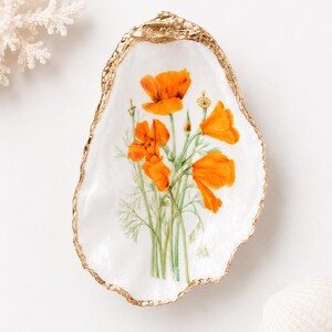 May include: A white oyster shell dish featuring a botanical illustration of bright orange California poppies with green stems and foliage. The edges of the shell are finished with a metallic gold trim, creating a decorative accent piece.