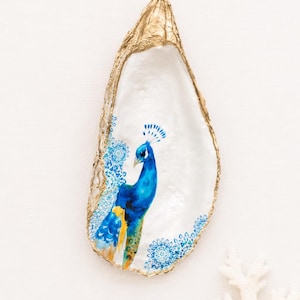 May include: A decorative oyster shell featuring a hand-painted blue and yellow peacock illustration on the interior. The shell has a smooth white pearlescent surface and a metallic gold painted edge, accented with small blue floral mandala patterns.