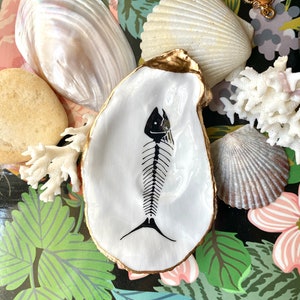 Puede incluir: Un plato de concha de ostra blanca con un dise&ntilde;o de esqueleto de pez pintado a mano en negro en el centro y un borde pintado en oro met&aacute;lico. La concha est&aacute; rodeada por varias conchas naturales, un trozo de coral blanco y una cadena dorada.