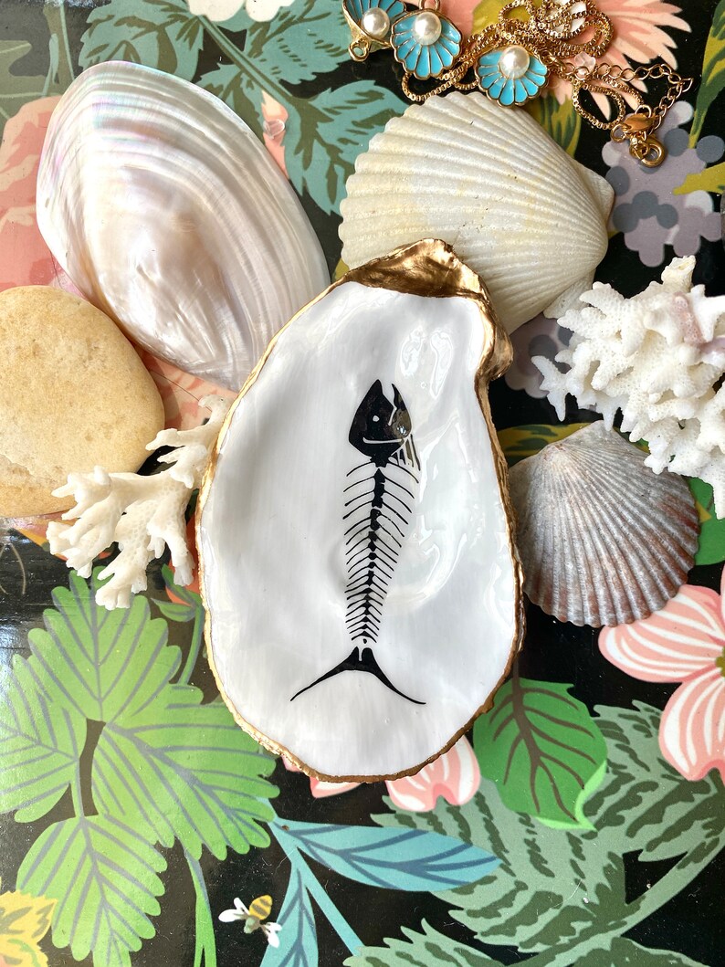 Puede incluir: Un plato de concha de ostra blanca con un dise&ntilde;o de esqueleto de pez pintado a mano en negro en el centro y un borde pintado en oro met&aacute;lico. La concha est&aacute; rodeada por varias conchas naturales, un trozo de coral blanco y una cadena dorada.