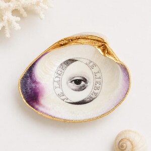 May include: A decorative clam shell dish with a gold-painted rim and a purple-tinted interior. The center features a black and white illustration of an eye surrounded by a circular band containing the text JE LÄNGER JE LIEBER.