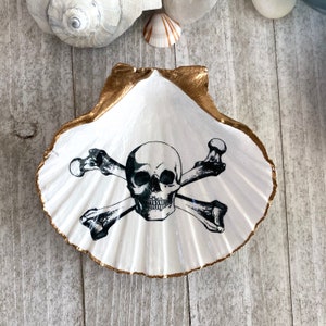 May include: A white seashell with a gold rim, featuring a black and white skull and crossbones design.