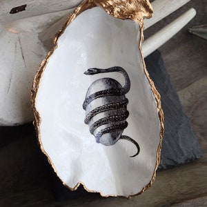 May include: A white ceramic oyster shell dish with a gold rim. The dish features a black and white illustration of a snake wrapped around an egg.