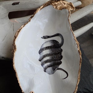 May include: A white ceramic oyster shell dish with a gold rim. The dish features a black and white illustration of a snake wrapped around an egg.