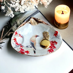 Puede incluir: Plato de cerámica blanca con forma de concha y borde dorado. El plato presenta un diseño floral rojo y blanco y una imagen pintada de un pájaro. Un collar de oro con un colgante redondo descansa sobre el plato.