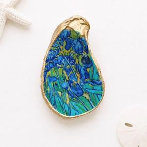 May include: An oyster shell featuring a painted reproduction of blue irises with green foliage. The edges of the shell are painted in a metallic gold finish. The shell is positioned between a white starfish and a white sand dollar.