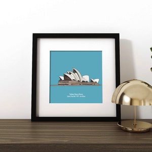 May include: A black framed print of a line drawing of the Sydney Opera House on a blue background. The print is on a wooden shelf with a gold lamp and a potted plant.