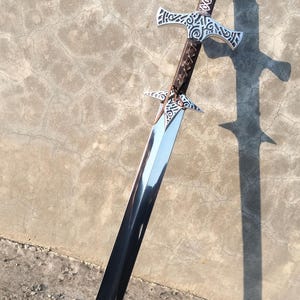May include: A silver and brown sword with intricate designs on the hilt and blade. The sword is lying on a gray concrete surface.
