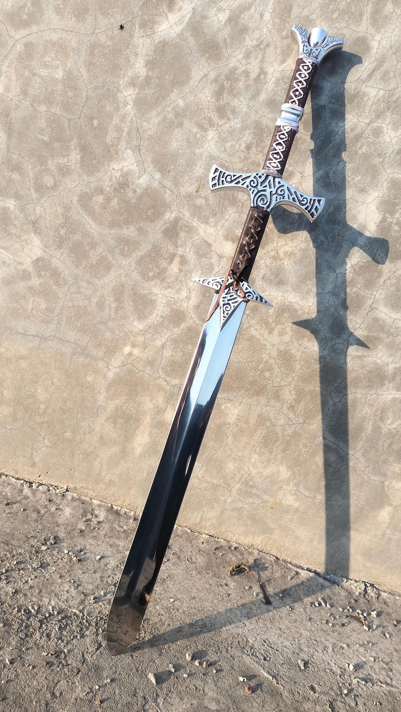 May include: A silver and brown sword with intricate designs on the hilt and blade. The sword is lying on a gray concrete surface.