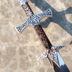 May include: A silver sword with a brown leather handle and intricate carvings. The sword has a double-edged blade and a crossguard with a decorative design.