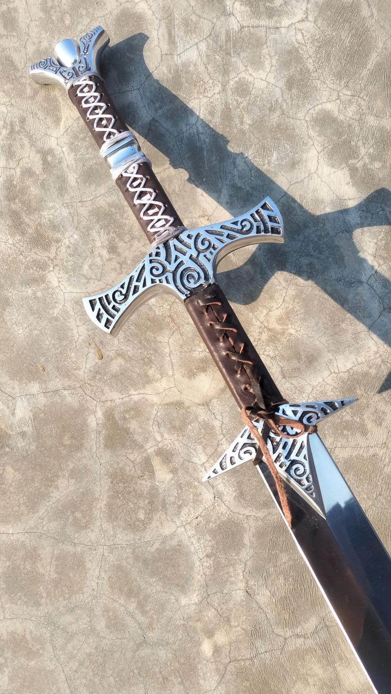 May include: A silver sword with a brown leather handle and intricate carvings. The sword has a double-edged blade and a crossguard with a decorative design.
