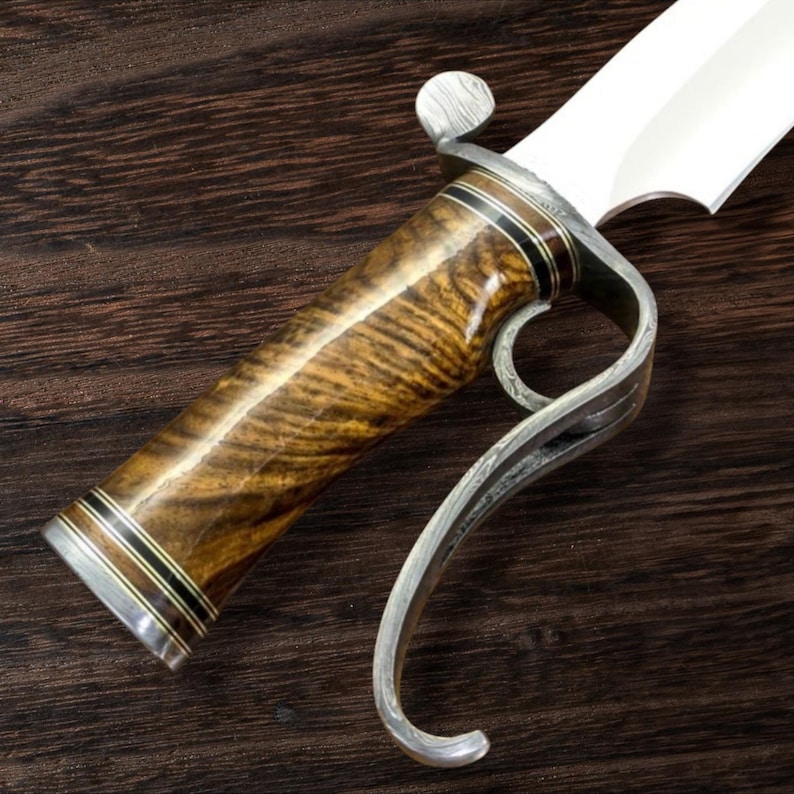 May include: Close-up of a custom knife with a wooden handle and a silver guard. The handle features a rich, brown wood grain with black and silver accents. The blade is bright and the guard has a curved design.