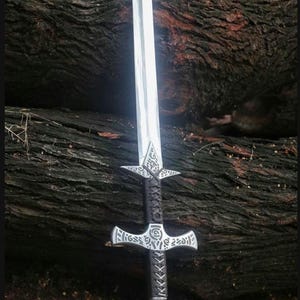 May include: A silver sword with a black leather handle and intricate metalwork. The sword has a cross guard and a pointed tip. The sword is resting on a wooden surface.