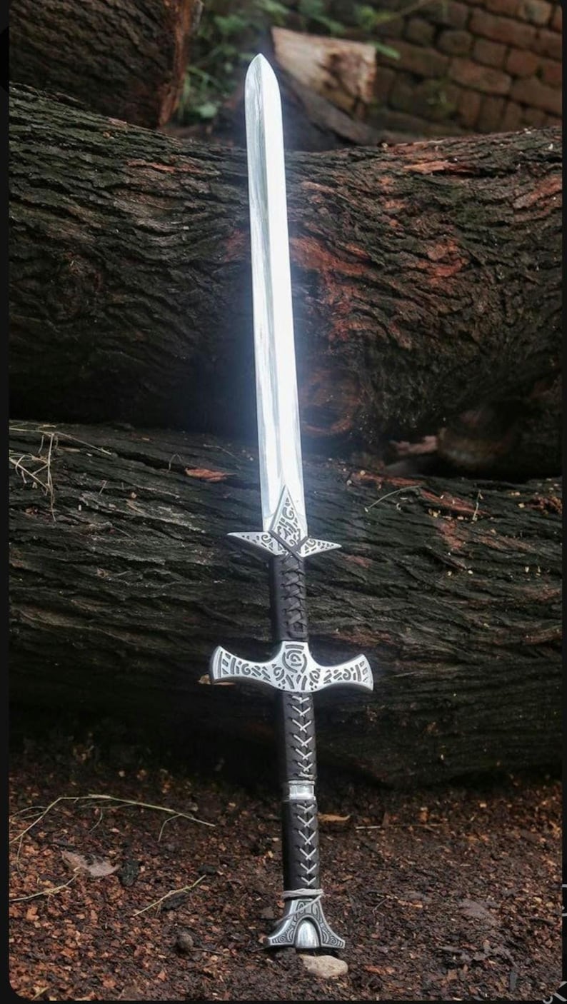 May include: A silver sword with a black leather handle and intricate metalwork. The sword has a cross guard and a pointed tip. The sword is resting on a wooden surface.