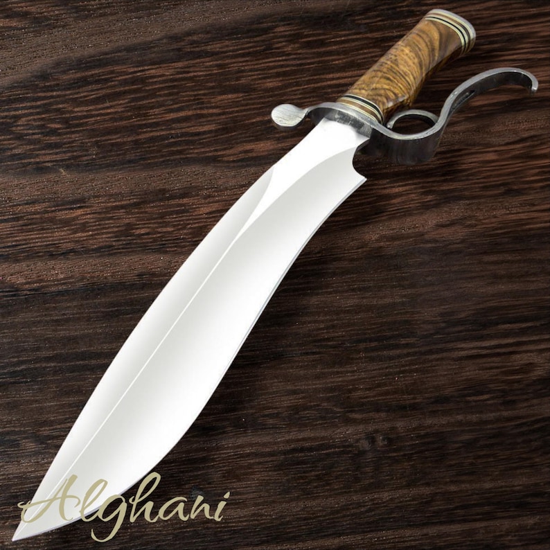 May include: A large, curved blade knife with a polished steel blade and a wooden handle. The handle is a light brown color with a textured grain. The knife has a metal guard and a curved handle. The word "Alghani" is written in cursive on the blade.