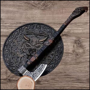Könnte beinhalten: Detaillierte Darstellung eines Wikingerbeils und -schilds vor einem Holzgrund. Das Beil weist aufwendige Schnitzereien und die Inschrift "WOLF MENTALITY PREVAILS" auf der Klinge auf. Das Schild zeigt ein Wolfskopf-Design.
