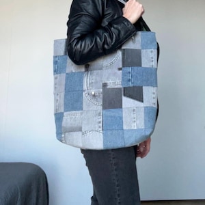 Peut inclure: Un sac fourre-tout en denim patchwork dans les tons de bleu et de gris. Le sac est composé de carrés de denim recyclé, avec des poches. Le sac a une forme rectangulaire avec un fond arrondi et une sangle gris clair.