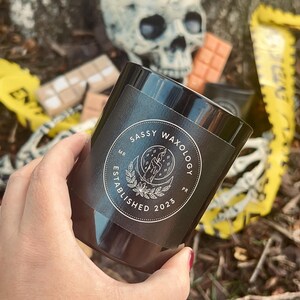 Könnte beinhalten: Ein schwarzes Kerzengefäß mit einem weißen Aufkleber, auf dem "Sassy Waxology" und "Gegründet 2023" steht. Der Aufkleber zeigt außerdem einen Mond und eine Hand, die eine Kerze hält.